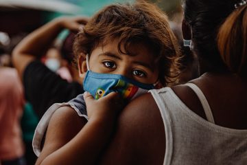 small child wearing mask, being carried in arms of mother